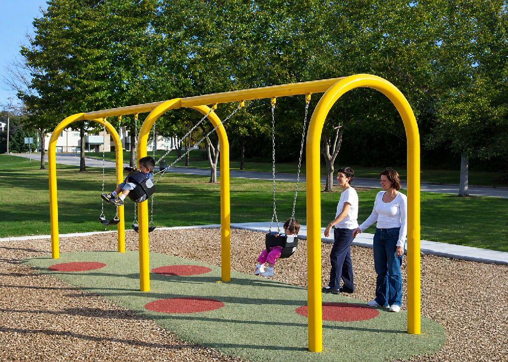 Young children swinging on a commercial playground swing set with bucket and belt seats while adults supervise at a public park playground.
